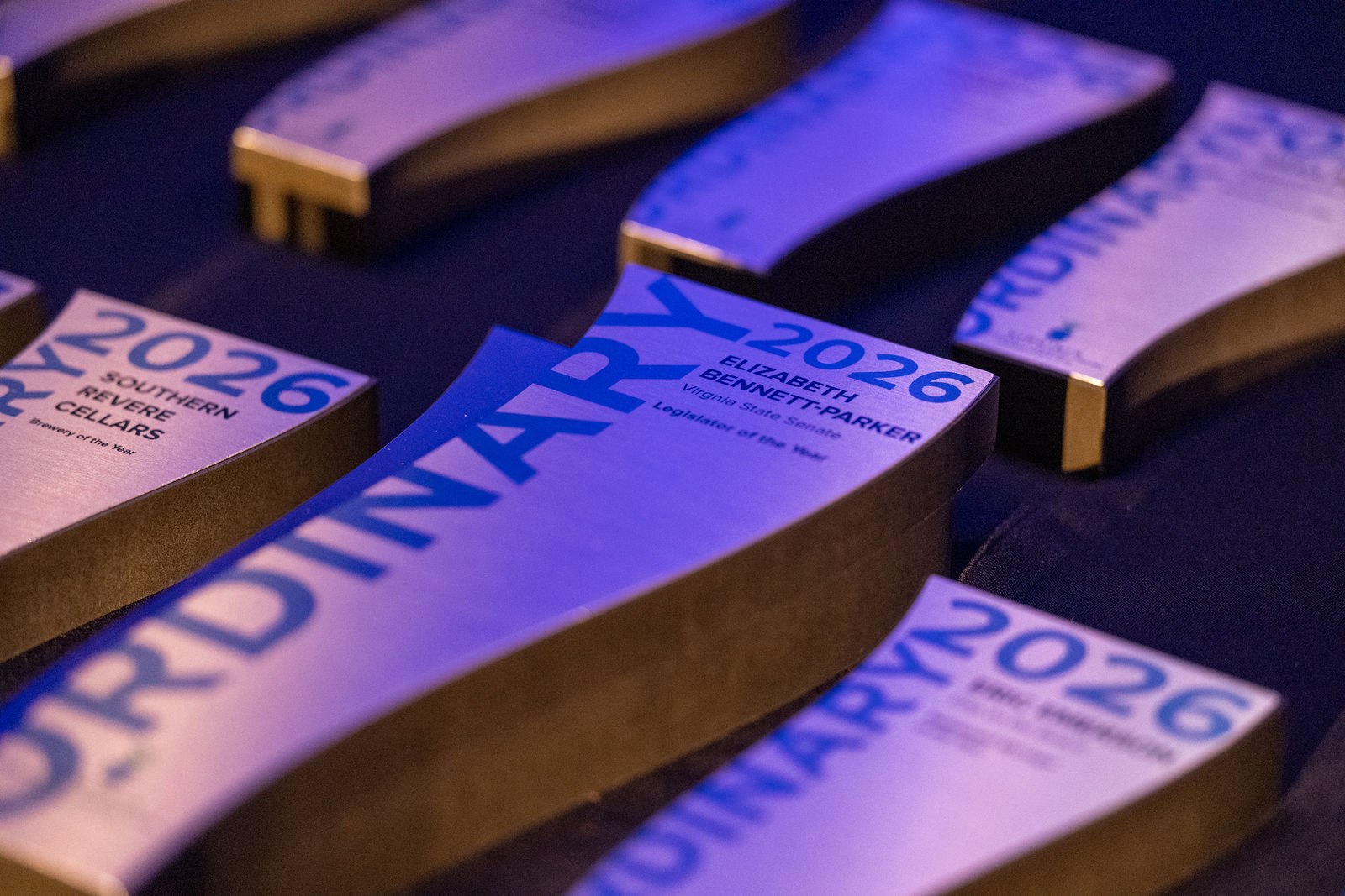Rows of 2026 Ordinary Award trophies laid out on a navy cloth under purple stage light. Each is a curved brushed-metal plate on a dark base reading ORDINARY 2026. The two in front are lettered for Southern Revere Cellars, Brewery of the Year, and Elizabeth Bennett-Parker of the Virginia State Senate, Legislator of the Year.