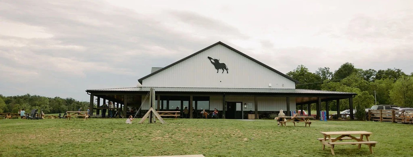 Chilly Hollow Brewing outside Berryville: a white gable-roofed barn with a black bull silhouette on the gable end and a covered porch wrapping the ground floor, with customers at tables under the roof and picnic tables spread across the mown lawn in front.