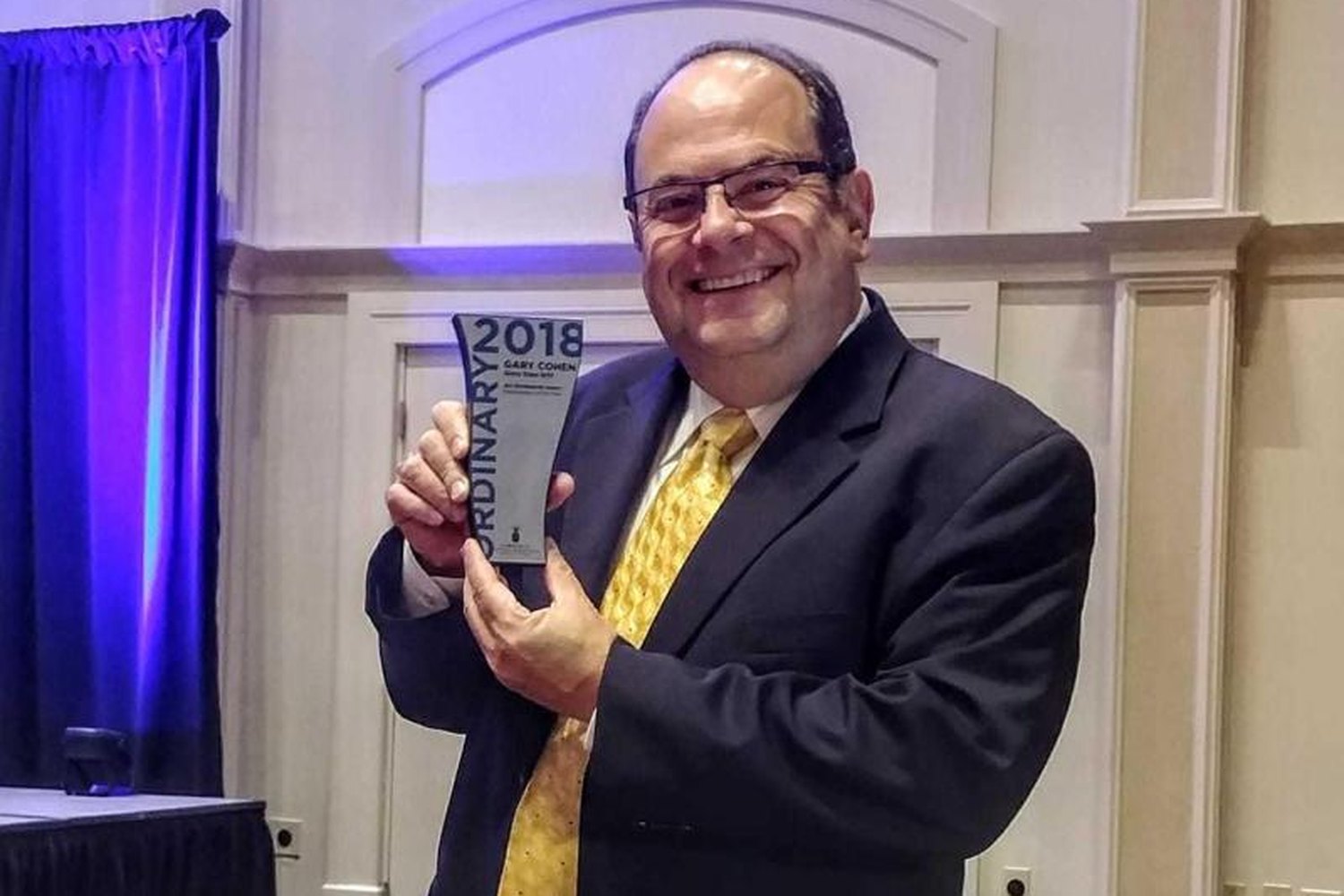 Gary Cohen in a dark suit and gold tie, smiling and holding up a glass Ordinary Award trophy engraved 2018, Gary Cohen, Glory Days Grill, in a hotel ballroom.