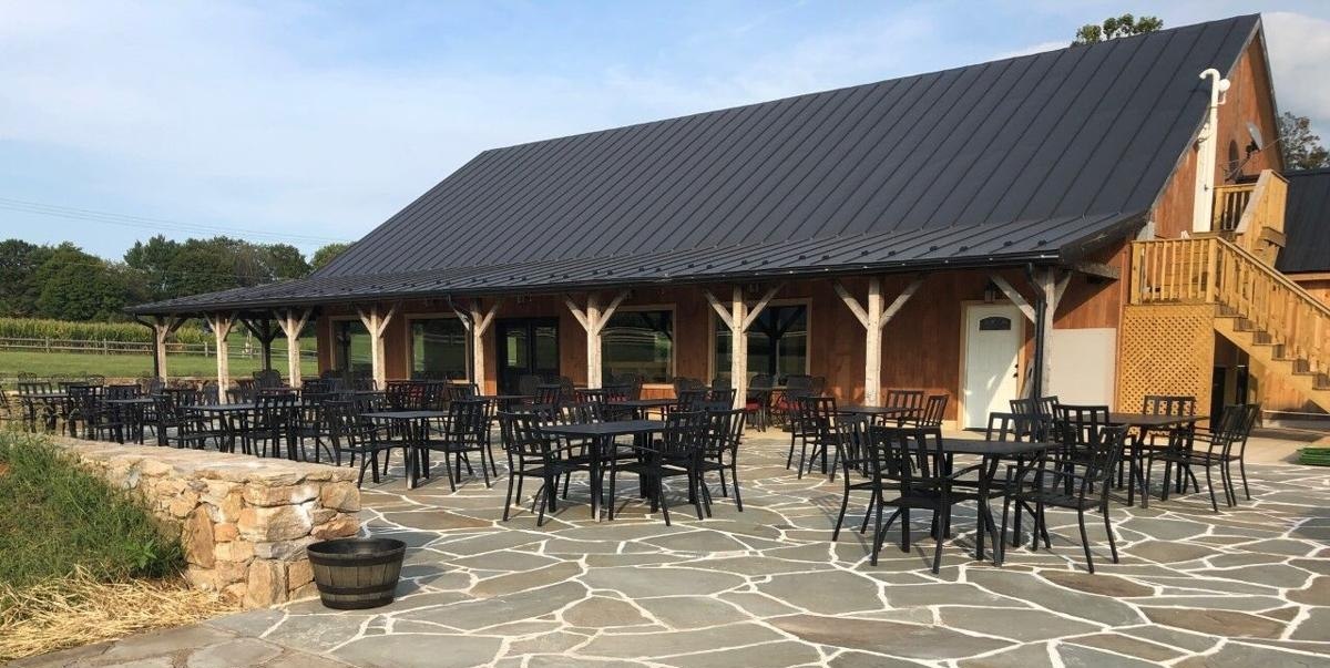 The timber frame taproom at Harvest Gap Brewery outside Hillsboro, with a covered porch on heavy posts, a black standing seam roof and a flagstone patio set with several dozen tables and chairs, a field of corn behind the fence line.