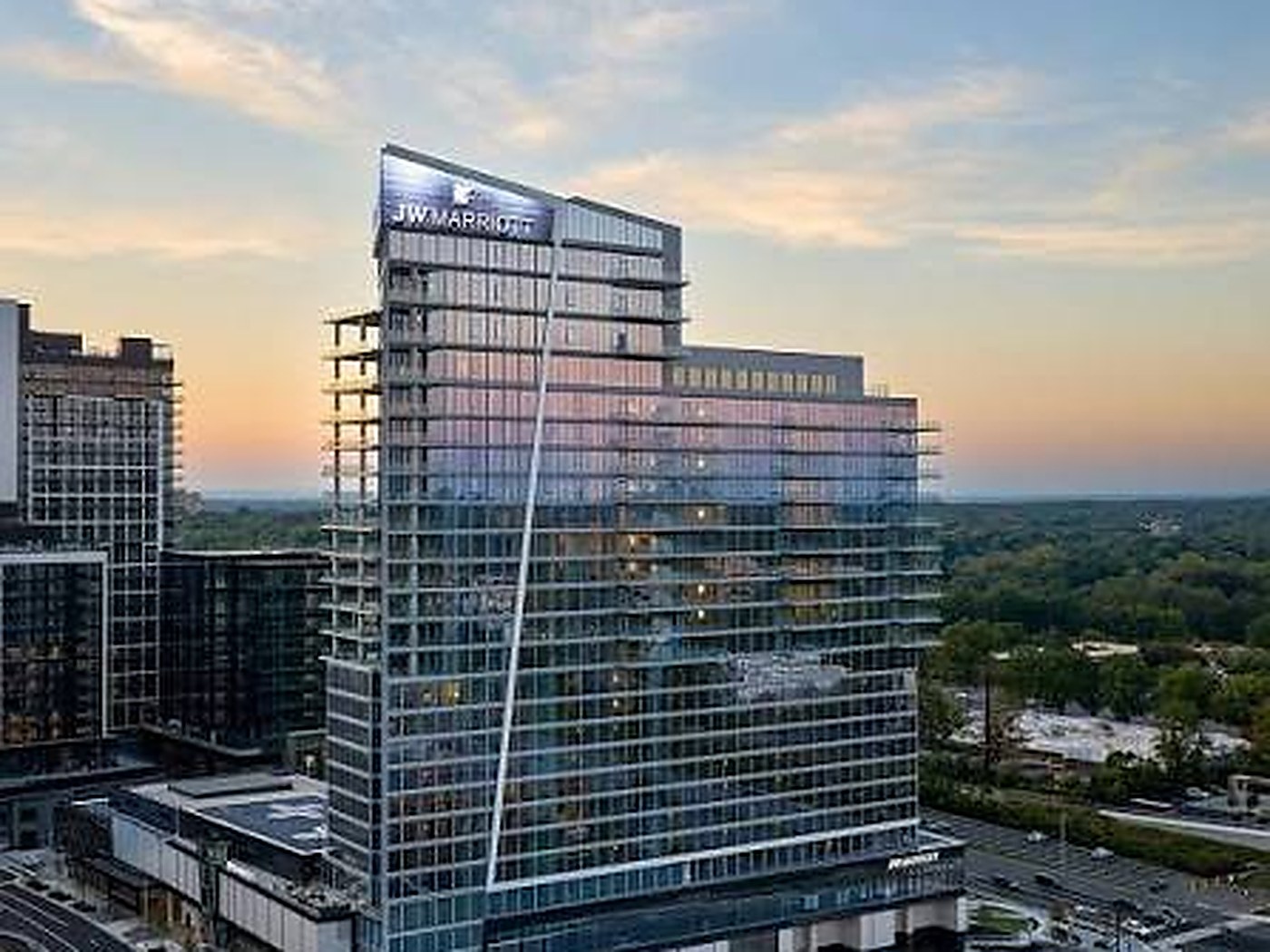 The JW Marriott Reston Station tower at dusk, a glass high-rise with lit rooftop JW Marriott signage, seen from the air with neighboring Reston Station buildings to its left and tree canopy and the Dulles corridor behind it.