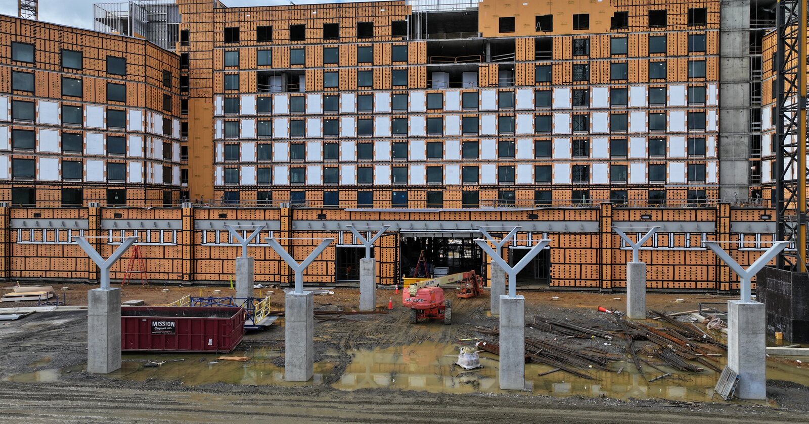 Aerial view of the Kalahari Spotsylvania construction site: a long multi-storey guest tower wrapped in orange sheathing behind a row of concrete columns and a muddy work yard.