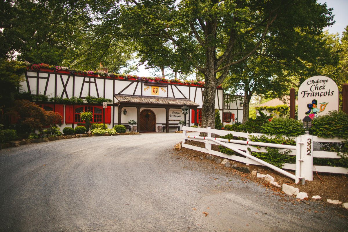 The half-timbered exterior of L'Auberge Chez Francois in Great Falls, with red shutters, flower boxes along the roofline and a white sign reading L'Auberge Chez Francois at the end of a gravel drive.