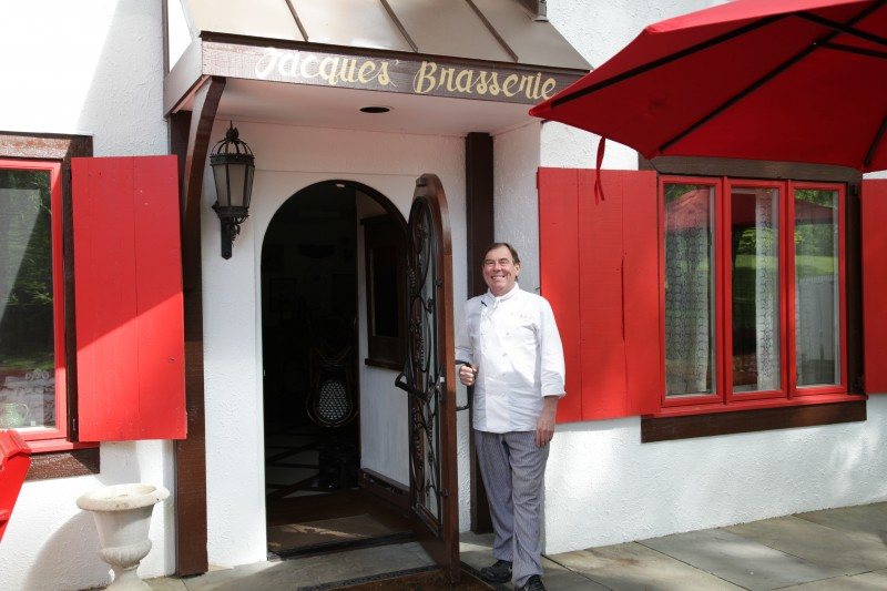 Chef Jacques Haeringer in white chef's whites, standing in the open doorway beneath the Jacques' Brasserie sign, flanked by red shutters and a red umbrella.