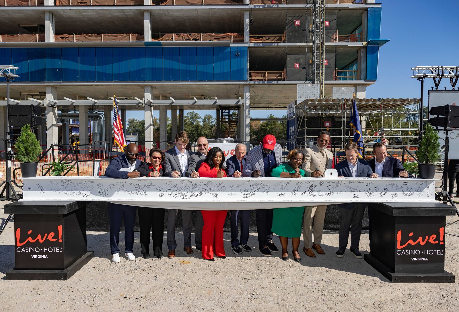 A dozen people in business clothes lean over a long white steel beam covered in signatures, signing it with markers. Two podiums carry the Live! Casino and Hotel Virginia logo, and the unfinished concrete and glass frame of the resort rises behind them under a clear sky.