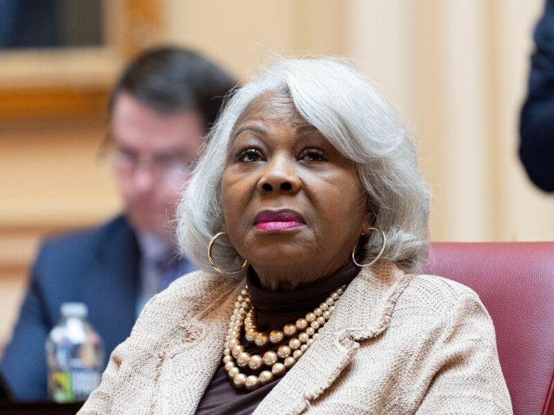 Virginia Sen. Louise Lucas seated on the Senate floor in a cream jacket and layered gold necklaces, listening with a neutral expression, another senator out of focus behind her.