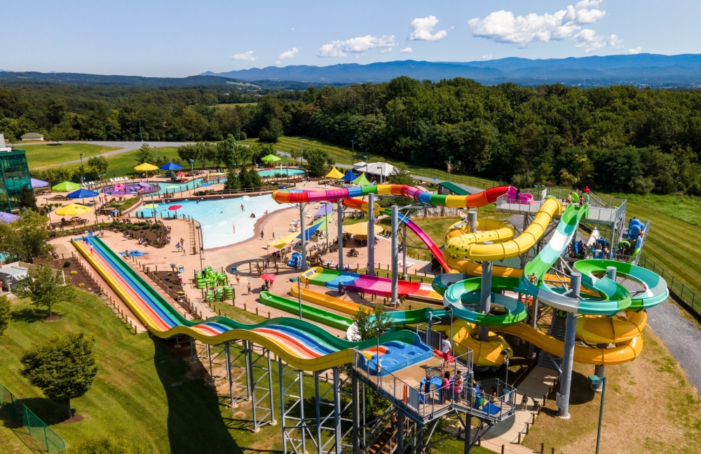 Aerial view of Massanutten Resort's outdoor water park: a wave pool, a row of racing slides in rainbow colours, and yellow and green tube slides on a tower, with the Blue Ridge mountains on the horizon.
