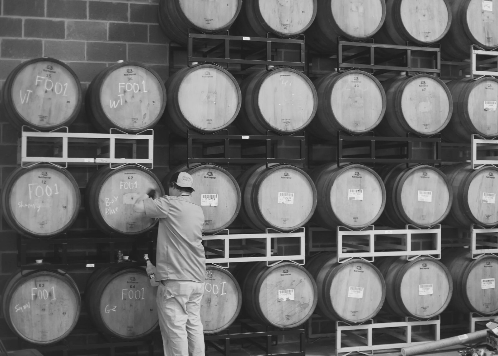 Black and white photograph of a brewery barrel room: rows of stacked oak barrels on steel racks, several chalk-marked with batch codes, as a worker in a cap reaches up to mark one of them.