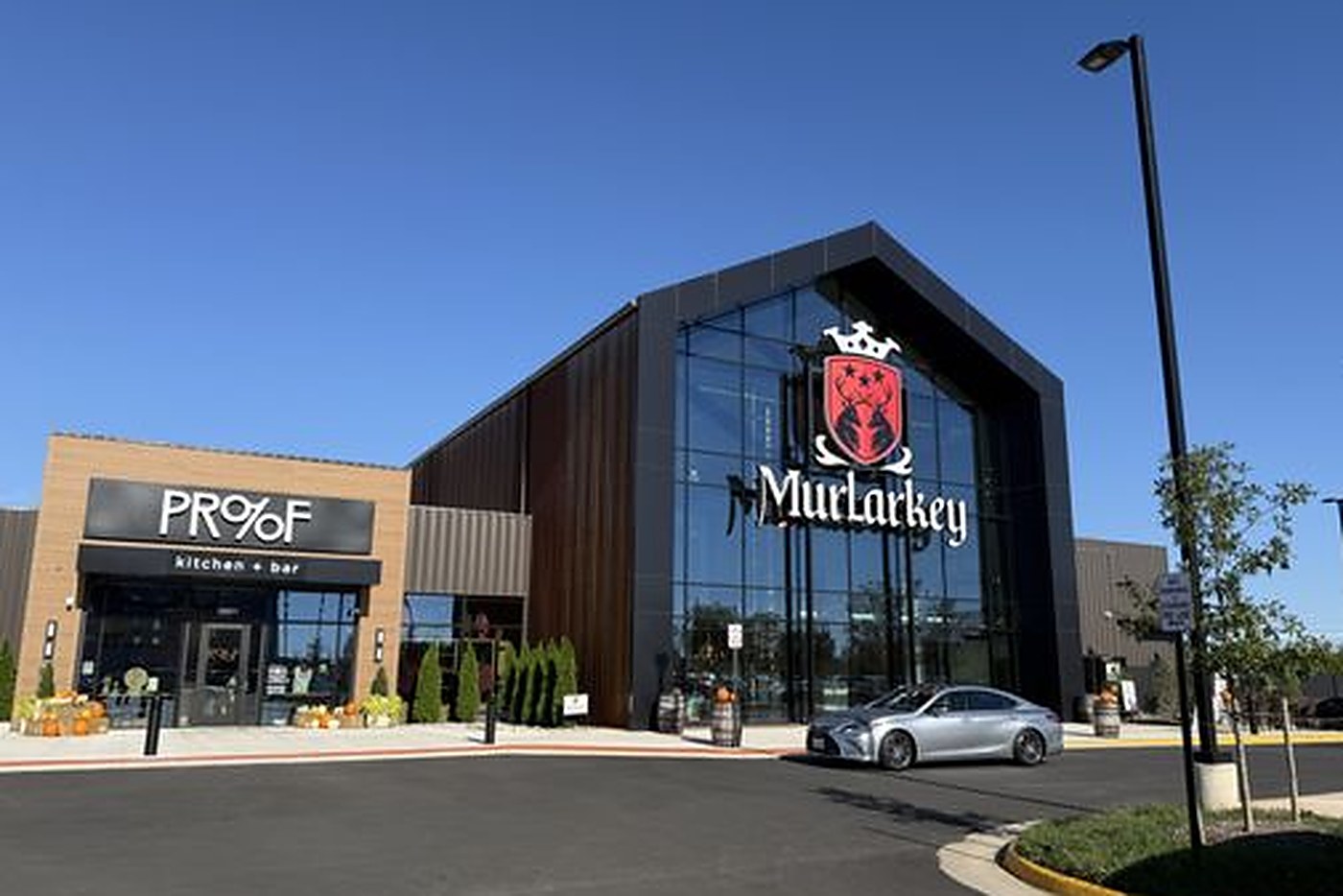 The MurLarkey Distilled Spirits building at NOVA Live in Manassas, a dark metal and timber structure with a tall glass front carrying the MurLarkey crest, seen from the parking lot on a clear day.