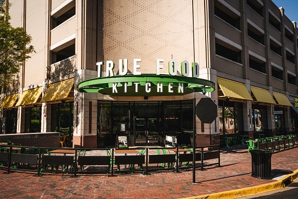 The corner entrance of True Food Kitchen at Reston Town Center, with a green oval sign above the glass doors, yellow awnings along two brick facades, and empty green patio tables on the brick sidewalk.
