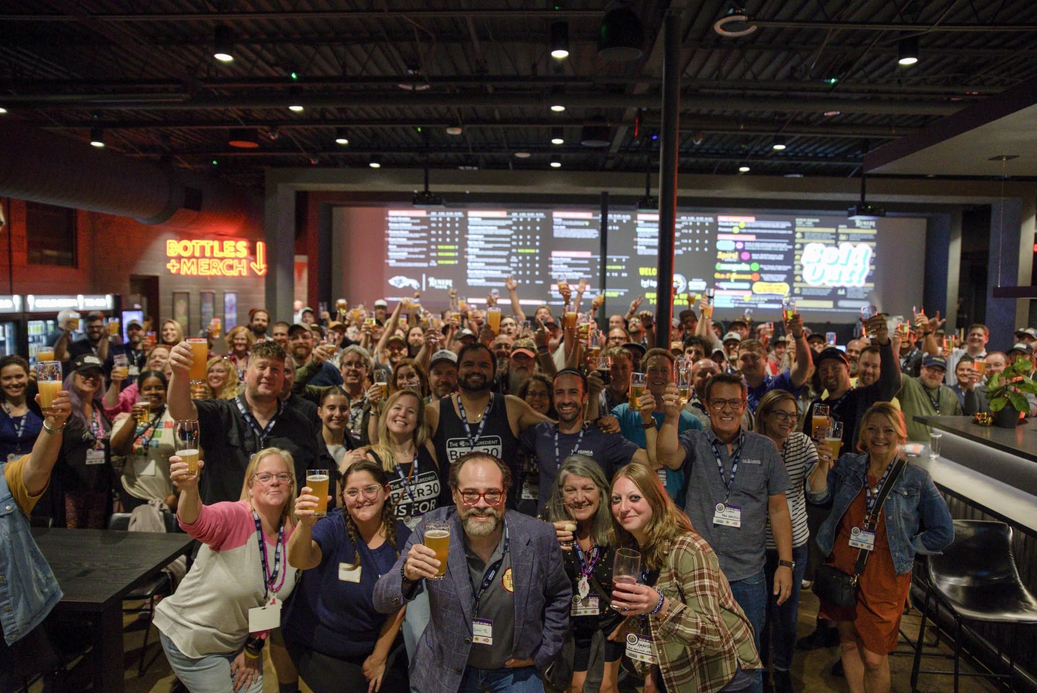 A large crowd of brewery professionals packed into a taproom, most of them grinning and raising a glass of beer toward the camera. Lanyards and name badges throughout, a neon Bottles and Merch sign on the left wall and a lit draft list behind them.
