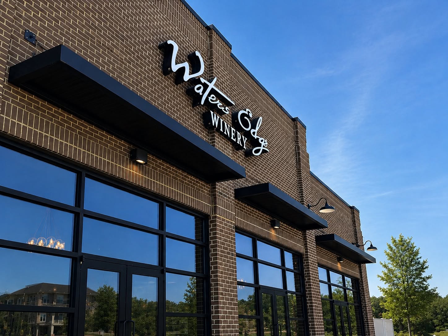 The brick storefront of Waters Edge Winery at dusk, with the script Waters Edge sign lit white above a row of tall black-framed windows and glass doors, and a chandelier visible inside.